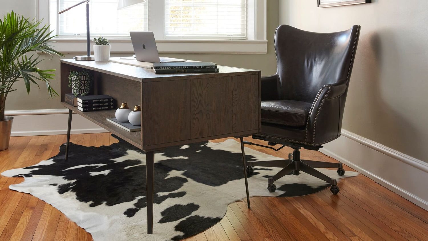 A modern home office with a dark wood desk on a black and white cowhide rug. The desk features minimalistic decor with potted plants, books, and a laptop. A black leather office chair with nailhead trim is situated behind the desk. Light streams through a window.