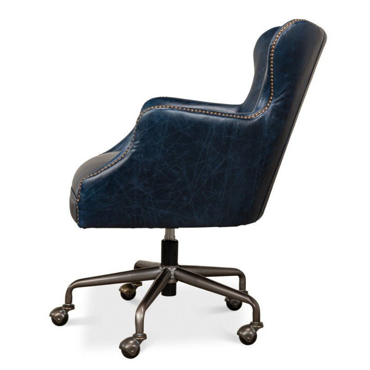 The Andrew Jackson Desk Chair in Chateau Blue by Sarreid showcases a side view with its high backrest and armrests. It is upholstered in pure aniline top-grade cowhide leather, accented with decorative nailhead trim along the edges, and rests on a black five-star base with caster wheels.