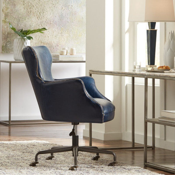 A modern home office showcases the Chateau Blue Andrew Jackson Desk Chair by Sarreid, crafted from pure aniline top-grade cowhide leather with silver rivet detailing. The chair is placed at a sleek metal desk adorned with a white lamp, a glass of water, and a bowl of snacks. Minimalist wall art and a potted plant complete the stylish space.