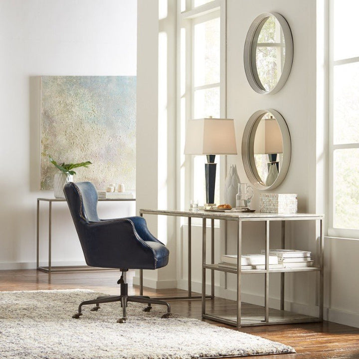 A modern home office features a Chateau Blue Andrew Jackson Desk Chair by Sarreid, complete with nailhead trim, in front of a glass-top metal desk. Two round mirrors and a table lamp sit on the desk, while abstract art and a small plant decorate the background. A rug covers the wooden floor.