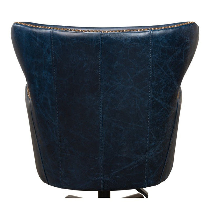 The Sarreid Andrew Jackson Desk Chair in Chateau Blue leather, viewed from behind, showcases a high, slightly curved backrest. Brass nailhead trim along the edges adds a touch of elegance to the chair. The pure aniline top-grade cowhide leather features a slightly wrinkled texture, giving the chair a vintage appearance.