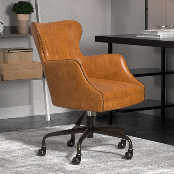 A modern office setting features the Andrew Jackson Desk Chair in Cuba Brown Leather by Sarreid, with a 5-ball caster swivel base, placed on a grey carpet. In the background are a white shelving unit adorned with a plant, books, and storage boxes, and a black desk with books and glasses on top.