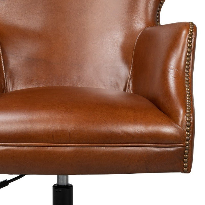 A close-up of the lower half of an Andrew Jackson Desk Chair in Havana Leather by Sarreid showcases its brown leather upholstery with nailhead trim on the edges. The chair, crafted in Vintage Havana leather, features a cushioned seat and is supported by a metal base with a height-adjustment mechanism visible at the bottom, embodying Transitional Design elegance.