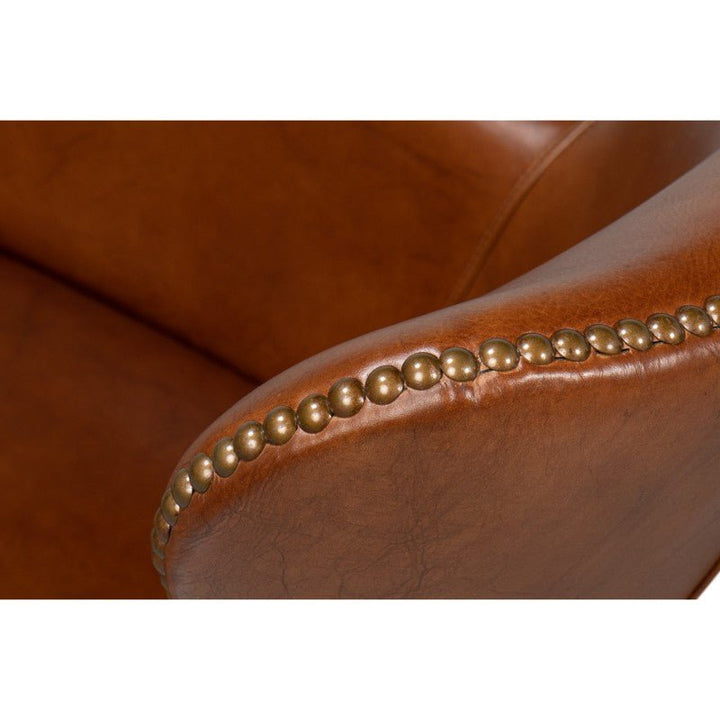 Close-up of the Sarreid Andrew Jackson Desk Chair in Havana leather's armrest. The armrest features decorative brass nailhead trim, adding a classic and sophisticated touch. The leather has a smooth texture with subtle, natural-looking creases and wrinkles, ideal for transitional design.