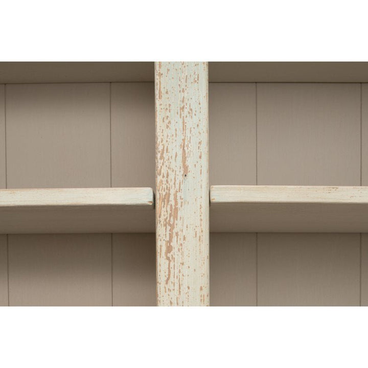 A close-up view of the Angelique Bookcase by Sarreid, showcasing its rustic, light green, distressed wooden design with a central vertical support. The shelves and backdrop feature a muted beige finish with visible wood grain that enhances its coastal charm and vintage aesthetic reminiscent of antique blue wash designs.