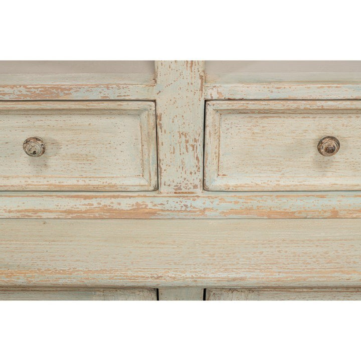 A close-up of the Angelique Bookcase by Sarreid reveals a weathered, rustic design crafted from reclaimed pine with an aged, distressed paint finish. The bookcase showcases two drawers adorned with vintage-style round metal knobs, embodying a shabby chic aesthetic. The wood’s visible grain and light, worn paint—with hints of pale blue—enhance its coastal charm.
