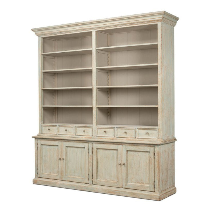 The Angelique Bookcase by Sarreid is a large, vintage-style wooden bookshelf with a distressed white finish, exuding coastal charm. It features multiple open shelves in the upper section divided into two columns, and a lower section with four cabinets and six small drawers, giving it a classic and rustic appearance.