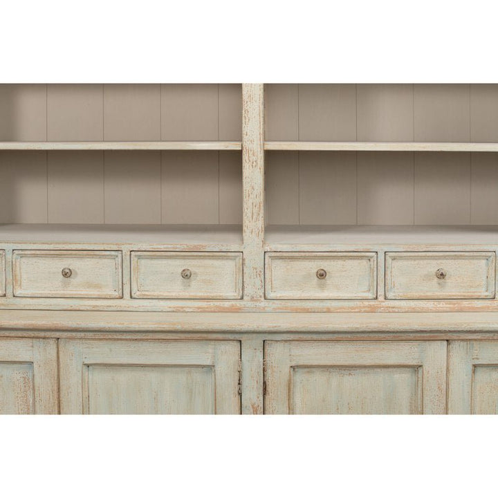 The Angelique Bookcase by Sarreid exudes coastal charm with its weathered wooden design. It features open shelves at the top, four small drawers in the middle, and wider cabinet doors at the bottom. Crafted from reclaimed pine, it boasts a distressed, vintage appearance with worn paint in a muted Antique Blue Wash hue.