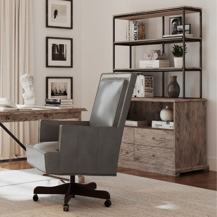 A home office with a wooden desk and a grey leather chair on wheels exudes rustic charm. Behind the desk, there's an Arcadian Lateral File Cabinet from Fairfield Chair and a wooden bookshelf adorned with books, decorative items, and a plant. The wall features framed black-and-white pictures, and a bust sculpture is placed on the desk. Light filters in from the left side.