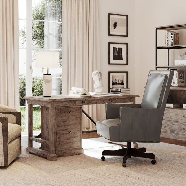 A home office featuring a burnished oak Arcadian Writing Desk and Mobile File by Fairfield Chair, adorned with a lamp and decorative bust, an ergonomic leather swivel chair, and a wooden bookcase holding books and décor. Large windows with beige curtains let in natural light, illuminating the serene and well-organized space.