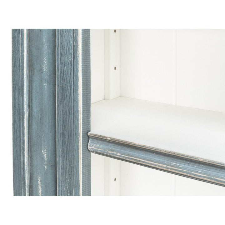 Close-up of a corner of the Banner Bungalow Bookcase by Sarreid, showcasing a distressed blue-gray finish with visible wood grain. The bookcase features white inner surfaces and a slightly protruding edge, highlighting the craftsmanship using Russian pine.