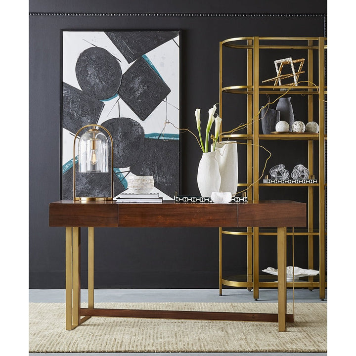 A contemporary room features a dark wooden console table with brass legs, complemented by the Elk Home Bell Jar 28'' High 1-Light Desk Lamp. A large abstract black and white painting and decorative items add flair, while a gold shelving unit adorns the dark wall. A beige rug elegantly covers the floor.