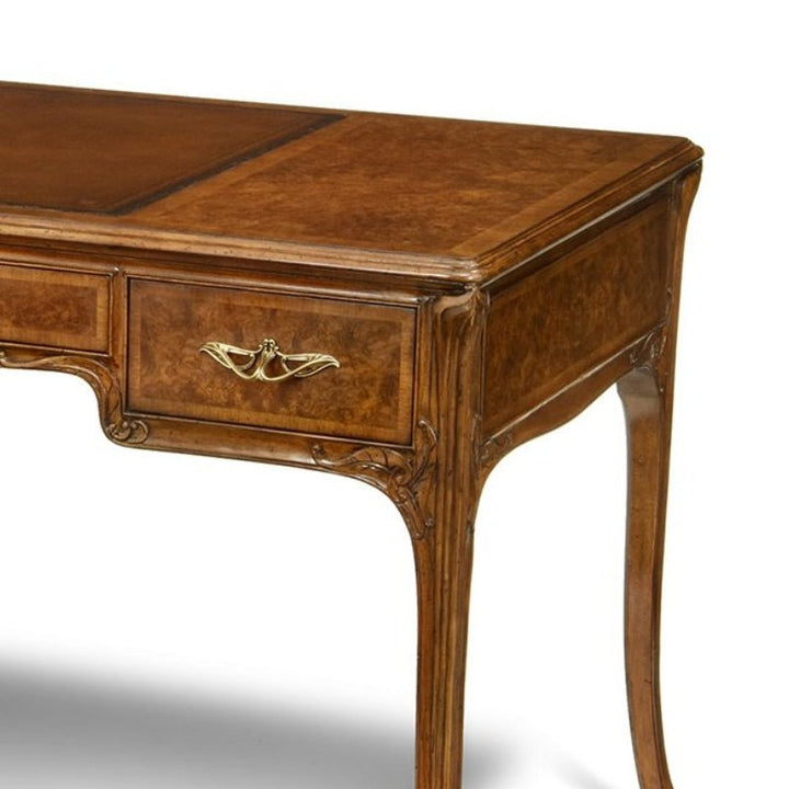 A close-up image of the elegant Aston Court Burl Walnut Lady's Writing Desk showcasing its detailed craftsmanship. The desk features a smooth, polished surface, a decorative brass handle on a shallow drawer, and intricately carved legs adorned with ornate patterns along the edges.