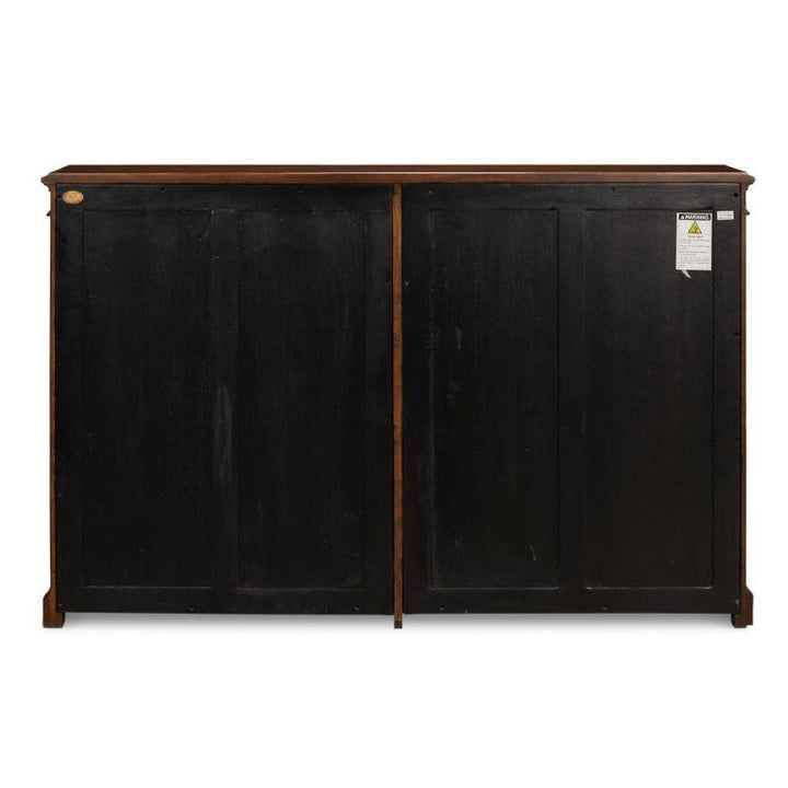 The image shows the back view of a Carmel-By-The-Sea Bookcase by Sarreid, featuring two large panels in a black wood finish. The top edge of the Walnut bookcase is adorned with antique brass accents and has a brown finish. A warning label is visible on the right side, and the bookcase appears to be placed against a white background.