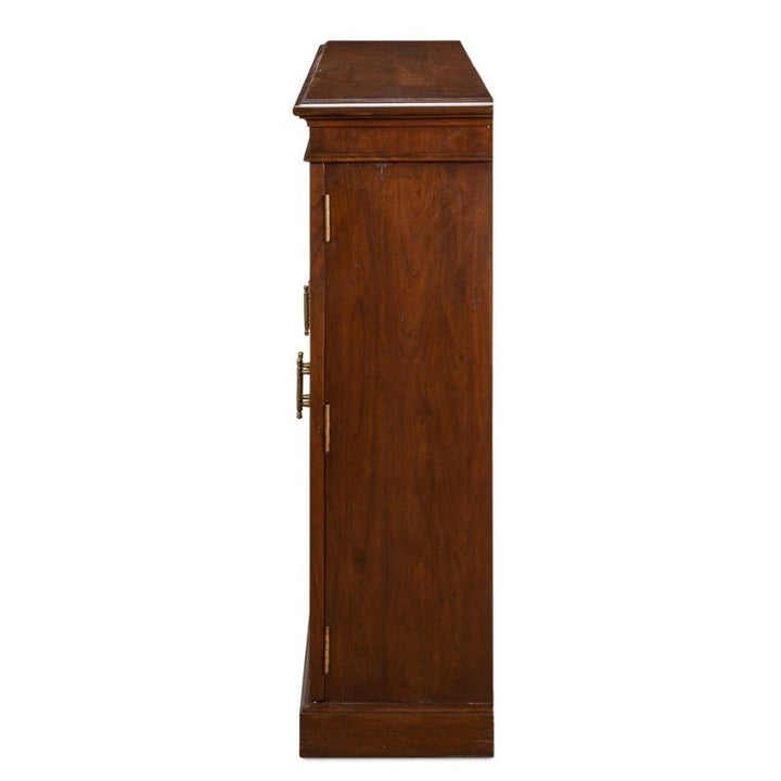 Side view of the Carmel-By-The-Sea Bookcase, Walnut by Sarreid. The solid walnut bookcase features one visible side adorned with two antique brass accents and a door handle. The polished brown finish enhances its elegant look, while the flat top surface slightly overhangs the front edge. Designed to sit directly on the floor, its base is solid and sturdy.