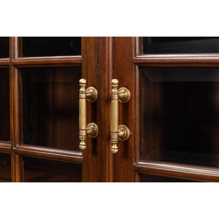 Close-up of the Sarreid Carmel-By-The-Sea Bookcase in solid walnut, featuring glass panes and intricately designed antique brass accents. The cabinet doors are dark wood with cylindrical handles showcasing decorative detailing, mounted on circular backplates. The glass panes offer a glimpse inside the bookcase.