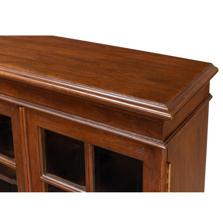 A close-up view of the Carmel-By-The-Sea Bookcase, Walnut by Sarreid showcases a polished solid walnut cabinet with a decorative top edge and glass-paneled doors. The wood boasts a rich brown finish, complemented by antique brass accents. This Sarreid bookcase appears sturdy and well-crafted, with the glass panels revealing the interior shelving.