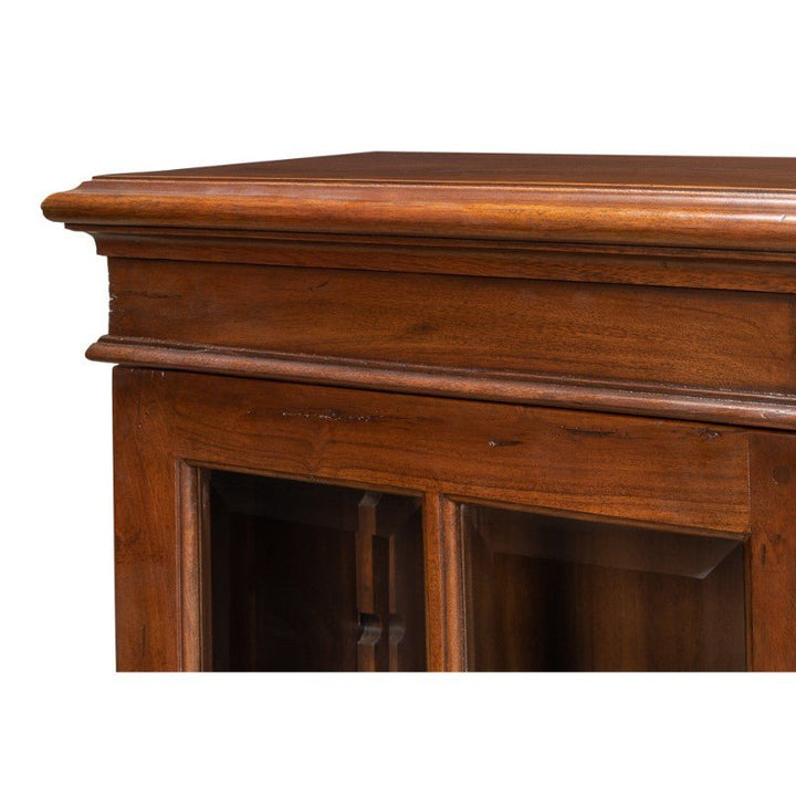 Close-up view of the corner of a Sarreid Carmel-By-The-Sea Bookcase, Walnut, with a polished finish. The bookcase features antique brass accents, a decorative edge, and glass panels on the door, showcasing its intricate craftsmanship and rich, warm color.