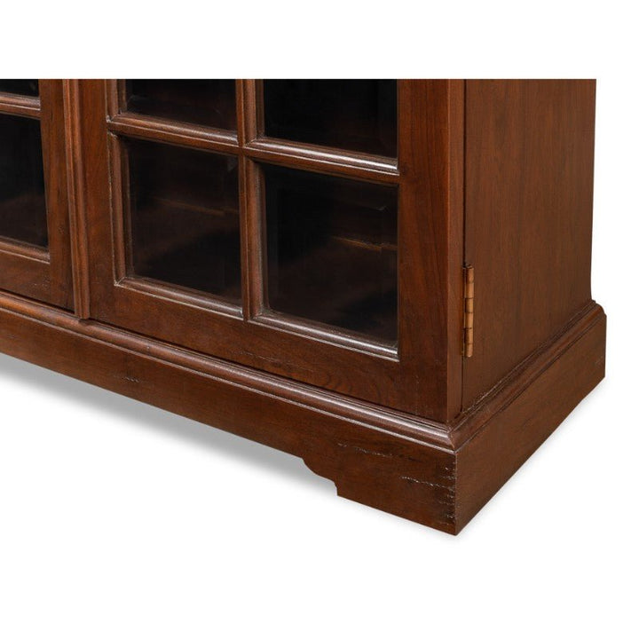 A close-up of the bottom section of the Carmel-By-The-Sea Bookcase by Sarreid shows its beautiful walnut design with a glass-paneled door. The solid walnut door is adorned with six glass panes and features antique brass accents, including a brass hinge. This exquisite bookcase has a rectangular base highlighted by slightly ornate molding at the edges, showcasing its rich brown finish.