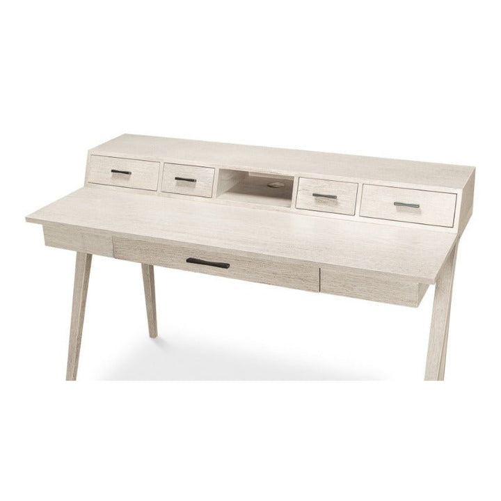 The Covet Desk by Sarreid is a light-colored wooden desk with an Ivory Bianca finish and minimalist design featuring angled legs. This modern desk offers a large primary workspace with one center drawer, complemented by an elevated section that includes four small drawers and a central open shelf. Sleek handles add to its practical functionality.