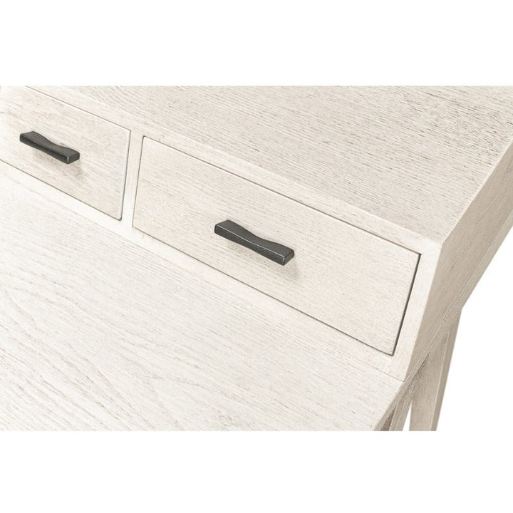 A close-up view of the Covet Desk by Sarreid showcases its light-colored wooden surface adorned with two small drawers at the top. The drawers are fitted with rectangular black handles. Enhanced by its Ivory Bianca finish that beautifully highlights the wood grain, this modern desk from Sarreid offers practical functionality paired with a minimalist design.