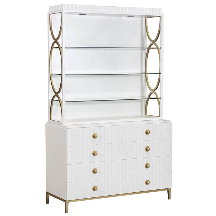 The East Camden Deck by Fairfield Chair is a stunning white mid-century modern cabinet characterized by a geometric frame and brass accents, sporting a pearl finish. It includes three glass shelves with three-way touch lighting overhead and a lower section with six drawers adorned with round brass handles. The piece stands proudly on slender brass legs.