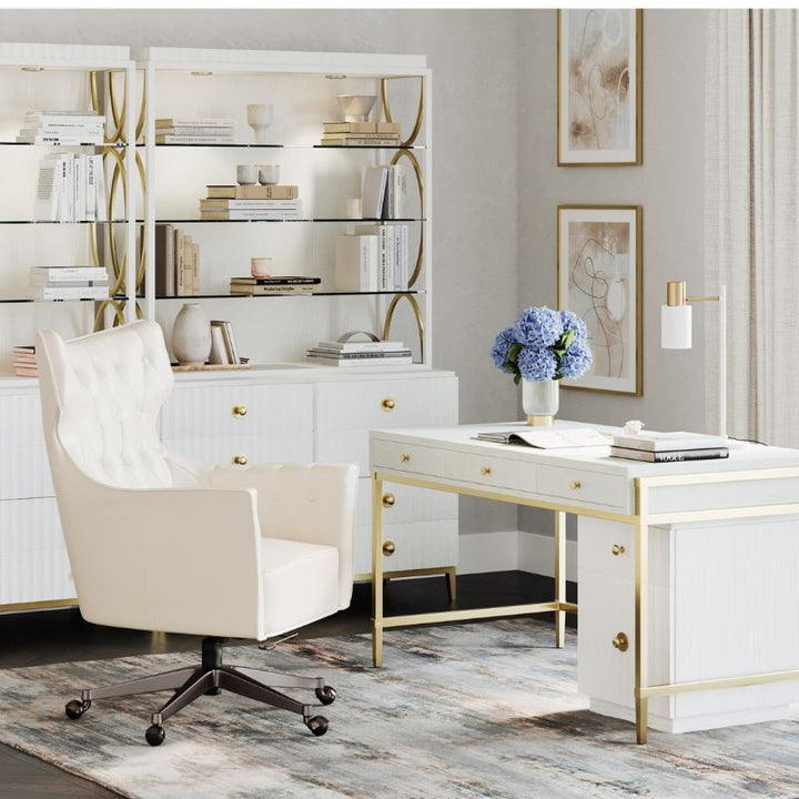 A modern home office with white and gold furniture. It features an East Camden Desk by Fairfield Chair with gold accents, a white tufted office chair, and a bookshelf holding decor items. The room is adorned with framed pictures on the wall, three-way touch lighting, and a vase of blue flowers on the desk.