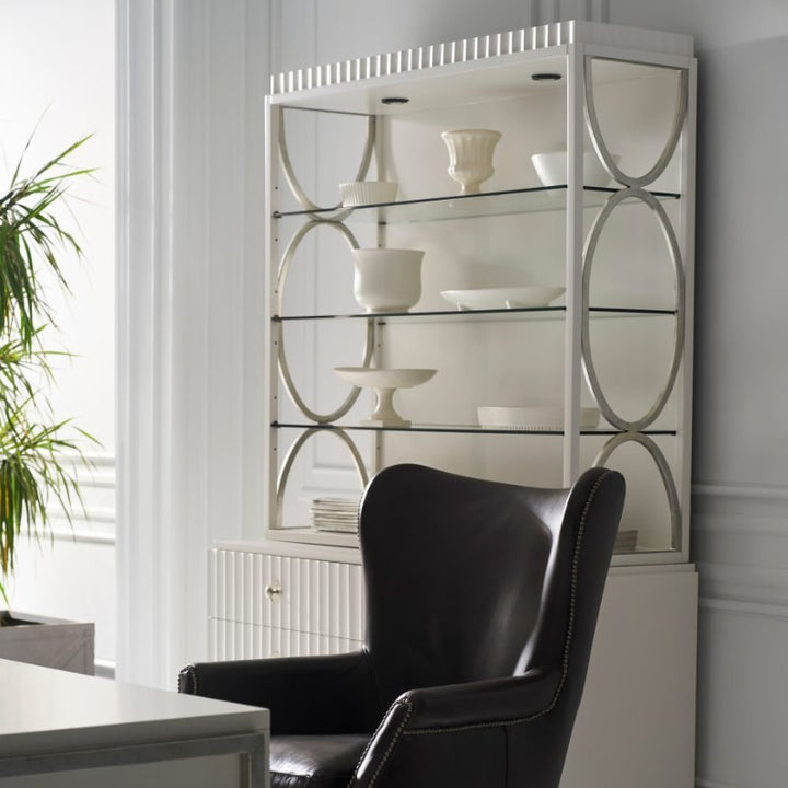 Against a light gray wall, the East Camden Deck by Fairfield Chair, a white shelving unit featuring a pearl finish and decorative circular patterns, displays an assortment of white porcelain items. Positioned in front of it is the Fairfield Chair Company's black leather chair with studded trim. On the left side, partially visible, is a green plant enhanced by three-way touch lighting.