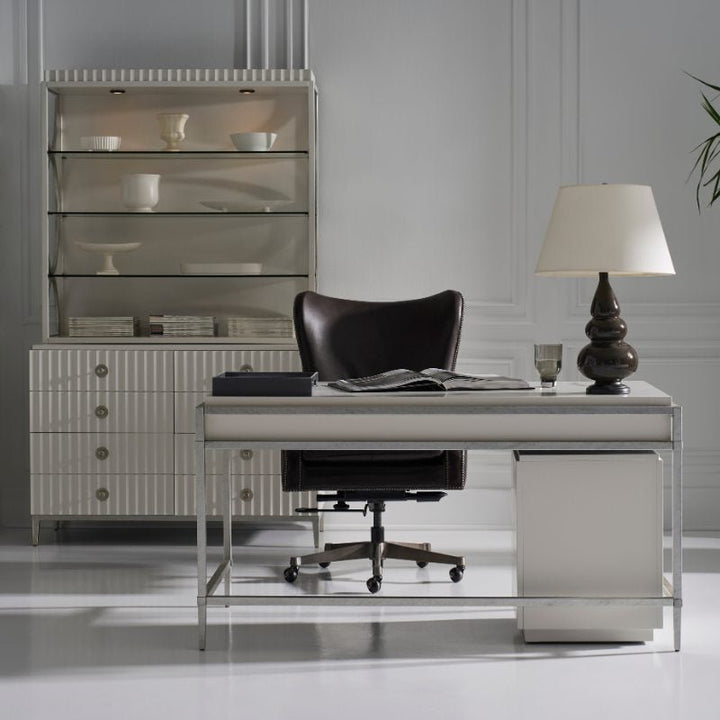 A modern, minimalist home office setup featuring the sleek white East Camden Desk by Fairfield Chair paired with a black swivel chair. The desk is adorned with a lamp, a glass of water, and a few books. Behind the desk is a white shelving unit displaying plates, bowls, and another stack of books. The desk's pearl finish complements the ambiance perfectly.