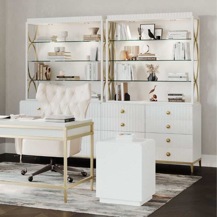 A sophisticated home office exudes casual elegance with a white desk featuring gold accents, paired with a cushioned chair. Behind the desk, two white shelves with Antique Brass hardware display books and decor. An East Camden Lateral File Cabinet by Fairfield Chair sits on a patterned rug, enhancing the room's charm.