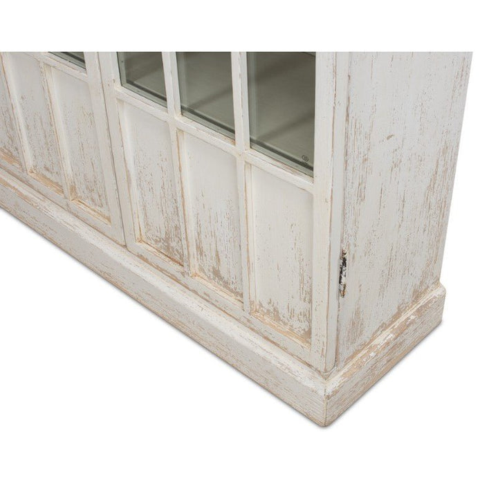 Close-up of the Edgar Allan Glass Bookcase by Sarreid, featuring distressed white wood and glass-paneled doors. The weathered finish exudes rustic charm reminiscent of a reclaimed pine bookcase. The photo captures the lower portion, including the base and part of the door hinges, highlighting its traditional design.