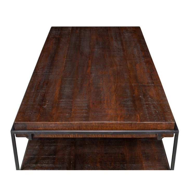 The Ernest Desk by Sarreid, crafted from mango wood and iron, features a rectangular surface with a dark brown rustic finish. The desk boasts a sleek black metal frame and includes a lower wooden shelf for additional storage or display. This piece could seamlessly suit an industrial desk setup, as showcased in an overhead photograph that highlights the rich texture of the wood.