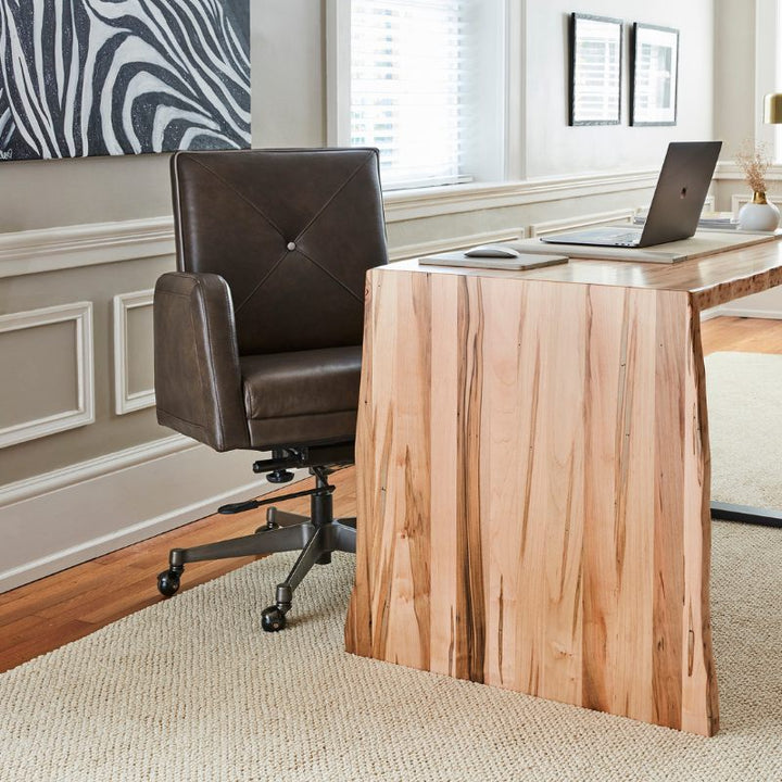 A modern home office with a sleek wooden desk and a Fairfield Chair Gherkin Office Swivel Chair made of dark brown aniline leather on a beige carpet. The desk features a laptop, a notebook, and a decorative plant. The room is well-lit with white framed windows and decorated with wall art, showcasing impeccable modern design.