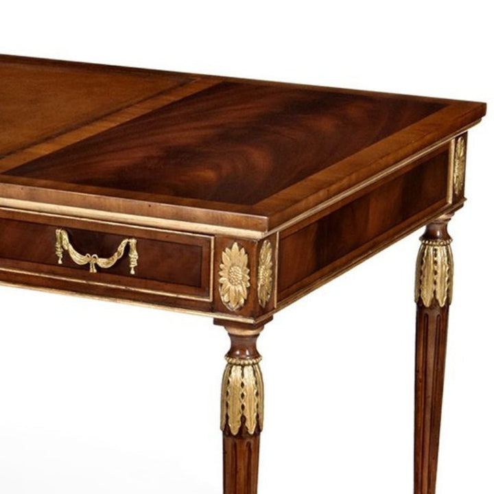 A close-up view of the Aston Court Gilt Mahogany Writing Desk reveals intricate gold embellishments and a decorative handle on the drawer. The desk's surface showcases a rich, polished wood grain, while the legs are adorned with ornate gold leaf accents, exemplifying luxurious craftsmanship.