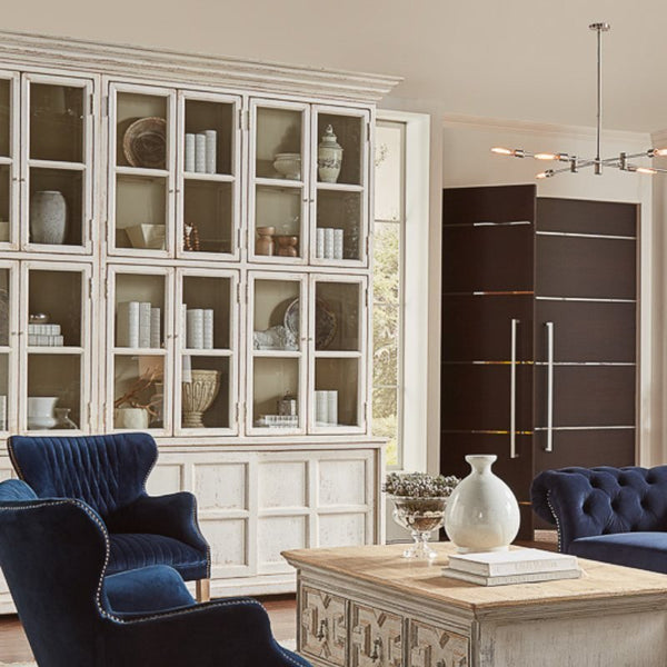 A sophisticated living room featuring two navy blue tufted armchairs, a Glass Front Bookcase from Sarreid filled with various decor items, a wooden coffee table adorned with a light decorative vase, and a modern light fixture hanging from the ceiling. The area exudes charm reminiscent of a farmhouse collection.