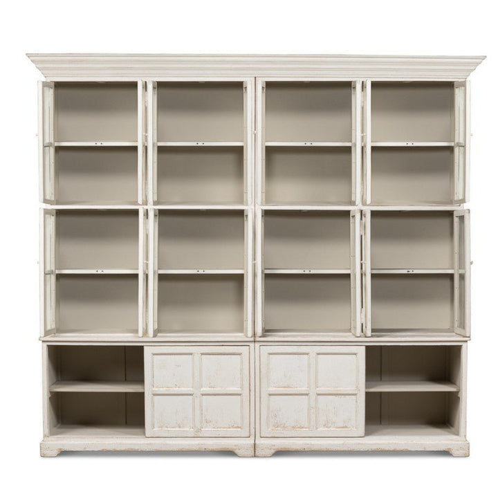 The Glass Front Bookcase - Sarreid by Sarreid is a large, white vintage-style unit featuring three rows of cubby shelves at the top and four cupboards with panel doors at the bottom. Part of our farmhouse collection, it boasts a distressed finish for a rustic, antique look. The shelves and cupboards are currently empty.