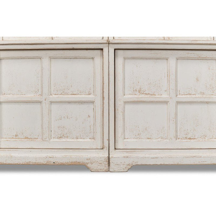 A close-up of the Glass Front Bookcase from Sarreid, showcasing its distressed white-finished wooden structure. This piece, part of the farmhouse collection, features two doors with six rectangular glass panels. The vintage aesthetic highlights worn textures and uneven paint, enhancing its rustic and antique charm for a delightful country-style addition to any home.