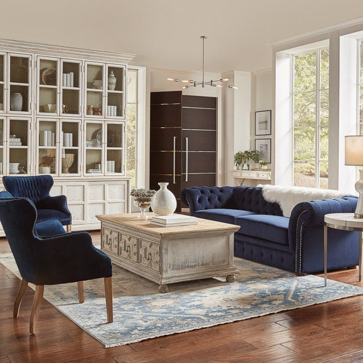 A chic living room featuring a navy blue tufted sofa with matching armchairs, a rustic wooden coffee table from the farmhouse collection, a blue patterned rug, and the large Sarreid Glass Front Bookcase. Natural light streams in through tall windows, brightening the space adorned with modern and cozy elements.