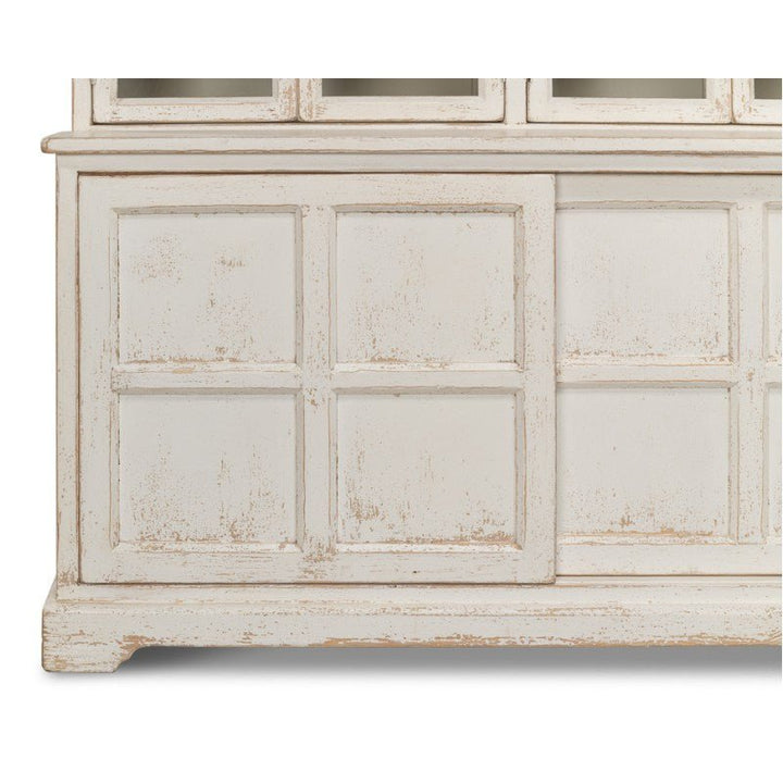 A close-up of the Sarreid Glass Front Bookcase reveals grid-like paneling with a rustic finish, showcasing a distressed white wooden design. The surface displays signs of wear and aging, enhancing its country-style vintage charm.