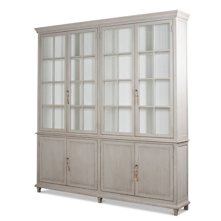 The Harper Glass Front Bookcase by Sarreid showcases an elegant design with a whitewashed finish, blending vintage charm with ample storage options. The upper section features glass-paneled doors that reveal interior shelving, while the lower section includes solid panel doors with handles for discreet storage. This cabinet stands gracefully on short legs.