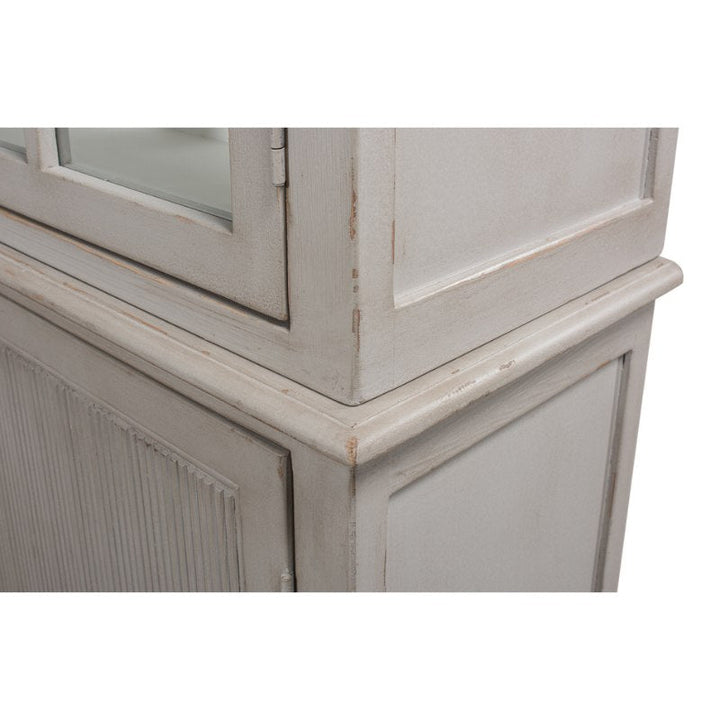 Close-up of a rustic wooden cabinet with distressed beige paint. The Harper Glass Front Bookcase by Sarreid features an elegant design, with a glass panel door on the upper section and vertical grooves on the lower section, highlighting its vintage style. It offers ample storage for your treasured items.