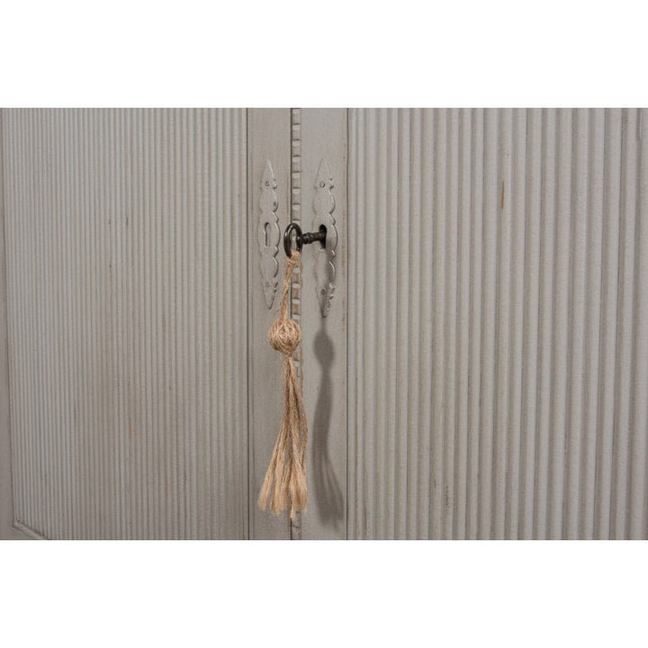 A close-up of a vintage-style white wooden door with a decorative keyhole and handle. Attached to the handle is a rustic key with a knotted rope tassel hanging from it. The door, reminiscent of the elegant design of Sarreid's Harper Glass Front Bookcase, has a ribbed texture. The overall look is shabby chic.