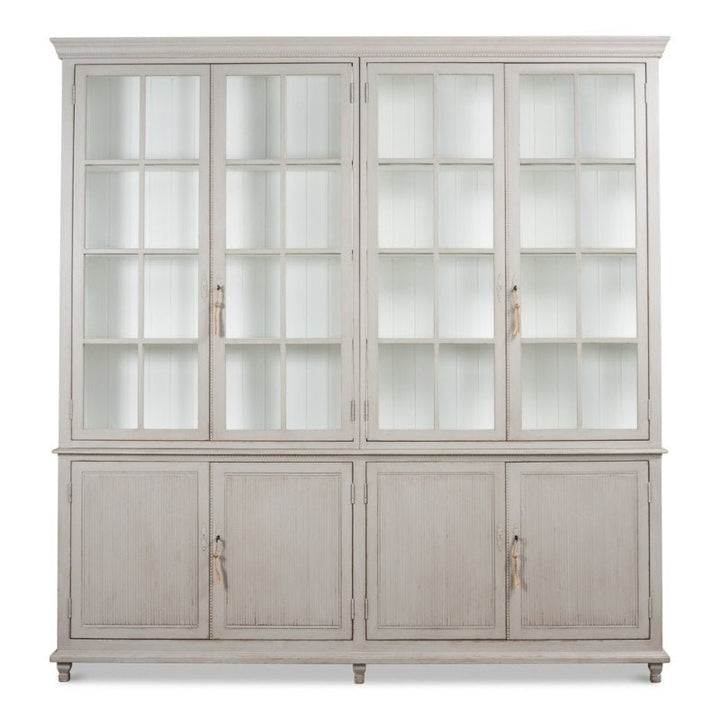 The Harper Glass Front Bookcase by Sarreid is an expansive, light grey wooden cabinet with a rustic finish and an elegant design. The top portion boasts four tall glass-paneled doors that reveal multiple shelves, while the bottom section includes four solid doors with minimalistic handles, providing ample storage space.