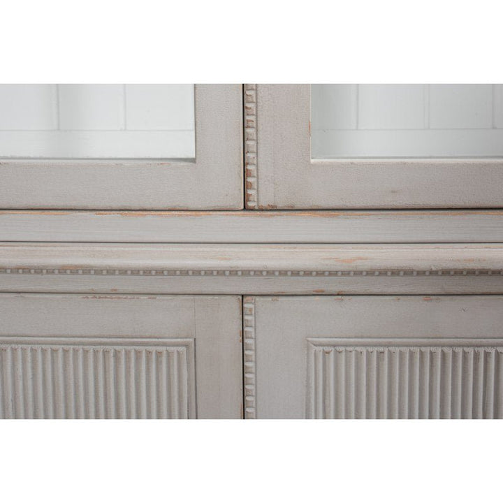 Close-up of the Harper Glass Front Bookcase by Sarreid, a distressed, vintage-style cabinet featuring glass panels and intricate decorative beading. It has vertical grooves on the lower panels and visible wear on its paint, indicating an aged patina. This elegant design is painted in an off-white or light gray color.