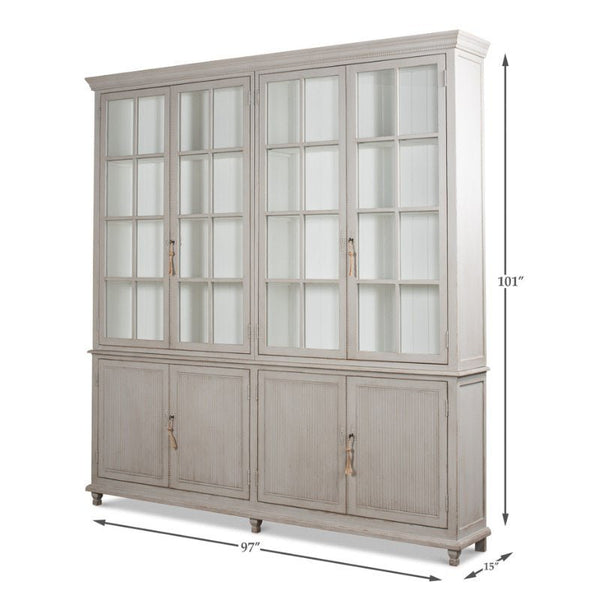 The Harper Glass Front Bookcase by Sarreid is a large, white wooden cabinet with an elegant design. It features glass-paneled double doors on the top section and solid double doors with metal handles on the bottom, providing ample storage. The cabinet stands 101 inches tall, 97 inches wide, and 15 inches deep.