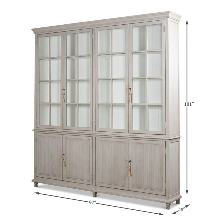 The Harper Glass Front Bookcase by Sarreid is a large, white wooden cabinet with an elegant design. It features glass-paneled double doors on the top section and solid double doors with metal handles on the bottom, providing ample storage. The cabinet stands 101 inches tall, 97 inches wide, and 15 inches deep.