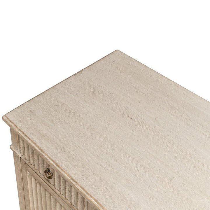 A close-up view of the top corner of a light-colored wooden cabinet. The cabinet, part of the Inspire Desk series by Sarreid, has a smooth surface and a vertical fluted design on the side panel, with a small round handle visible. The wood has a slightly weathered Stone Grey finish.