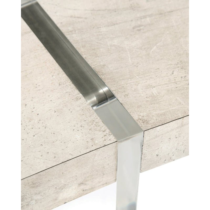 Close-up of a modern table corner with a concrete-like surface and sleek metal accents. The metal from the leg wraps around the edge and intersects the tabletop, reminiscent of the John Richard Loftus Desk 77" by John Richard, offering a rich tiza gesso finish that enhances its contemporary design and industrial feel.