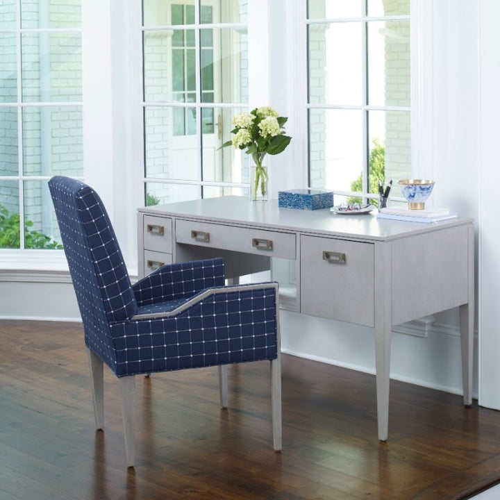 A home office setup featuring the Libby Langdon Patton Desk 54" by Fairfield Chair, showcasing a sheer dove gray finish with brass hardware and multiple drawers. Complementing the desk is a navy blue chair adorned with white grid patterns. The desk is accessorized with a vase of flowers, books, and stationery. Large windows in the background let in natural light, illuminating the wooden floor.