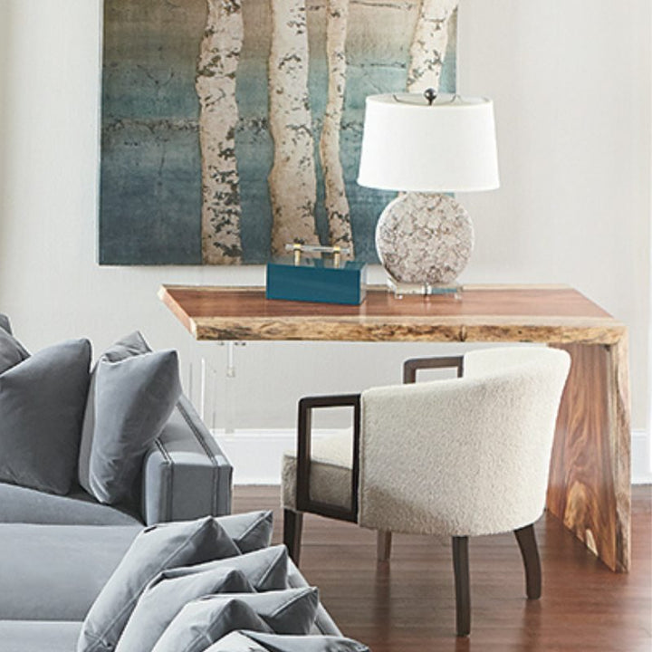 A cozy living room corner features a Live Edge Natural Guanacaste Waterfall Desk 54" with an acrylic base from Fairfield Chair, paired with a cushioned chair. A textured lamp and decorative box rest on the desk, showcasing refined craftsmanship. A large painting of birch trees hangs above, while a gray sofa is partially visible to the left.